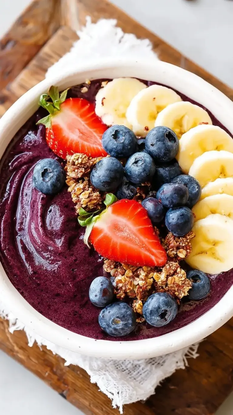 A vibrant purple acai bowl topped with fresh strawberries, banana slices, and crunchy granola in a white bowl.