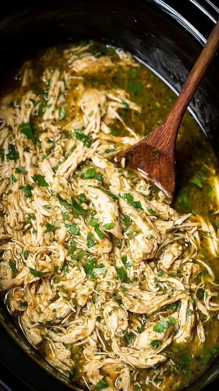 Shredded chicken in green salsa inside a slow cooker, garnished with fresh cilantro and lime